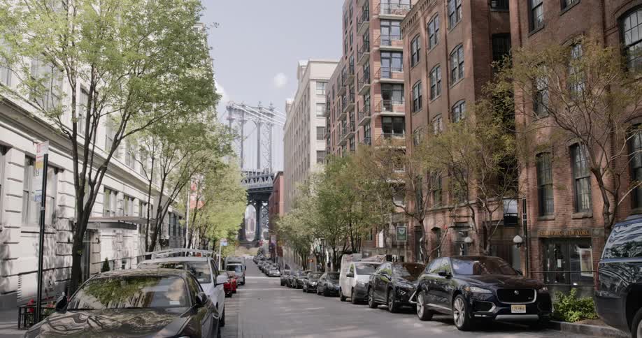 Cars are parked on both sides of the city street in front of apartment buildings with a bridge visible at the end