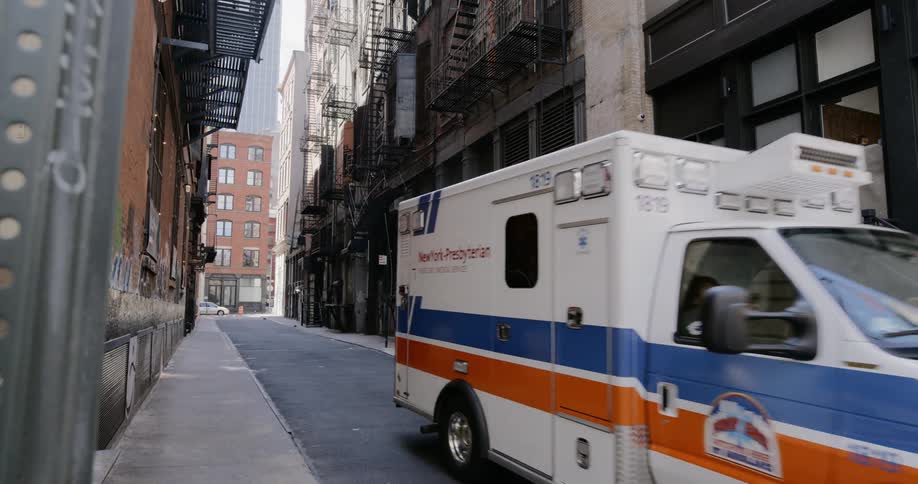 An ambulance slowly drives through an alley between buildings