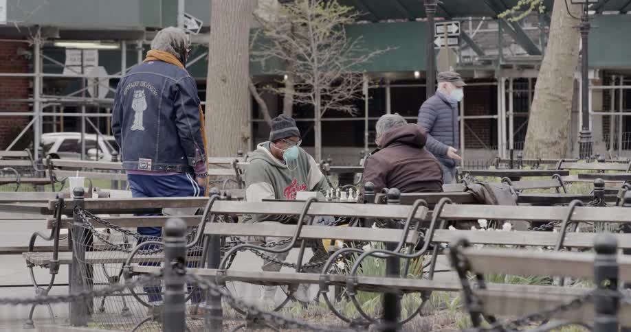 Two men sit outside and play chest as another man watches
