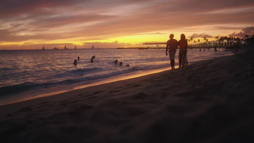 Couple has their arm around one another as they walk along the beach while the sun is setting and people are swimming in the ocean