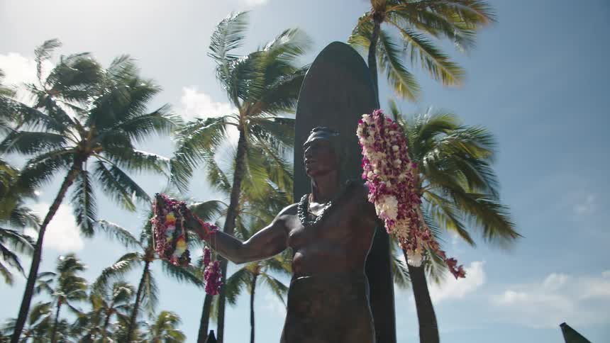Bronze statute of man against surfboard holding leis at a beach resort
