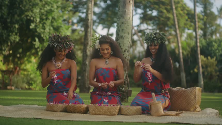 Three women sit and make leis