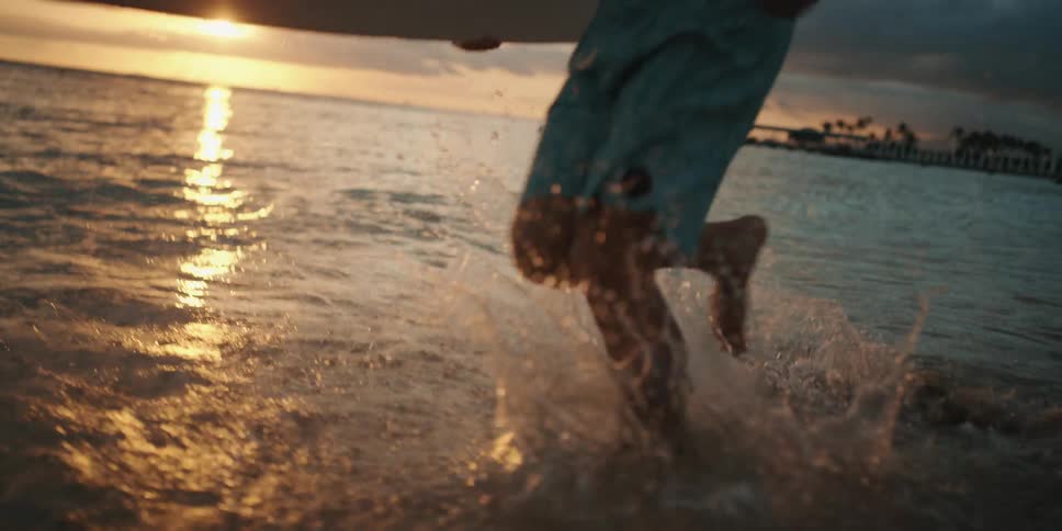 Man holding the surfboard runs along the beach at sunset next to the ocean