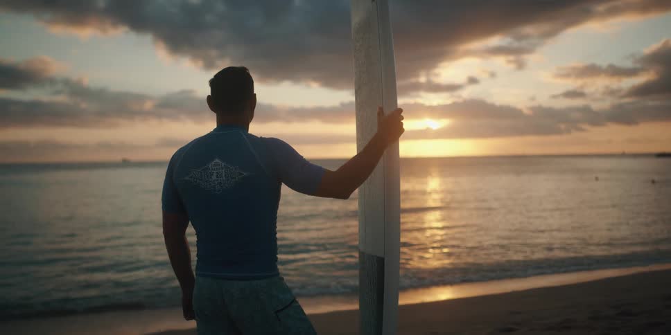 A man stands on the beach holding his board straight up in the sand and looking out at the ocean at sunset