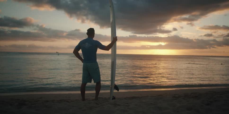 Man with surfboard stands on beach at sunset