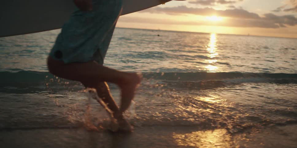 A surfer running along a beach at sunset carrying a surf board