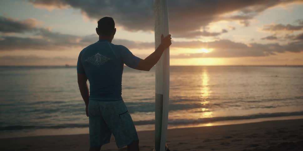 Man with surfboard stands on beach at sunset