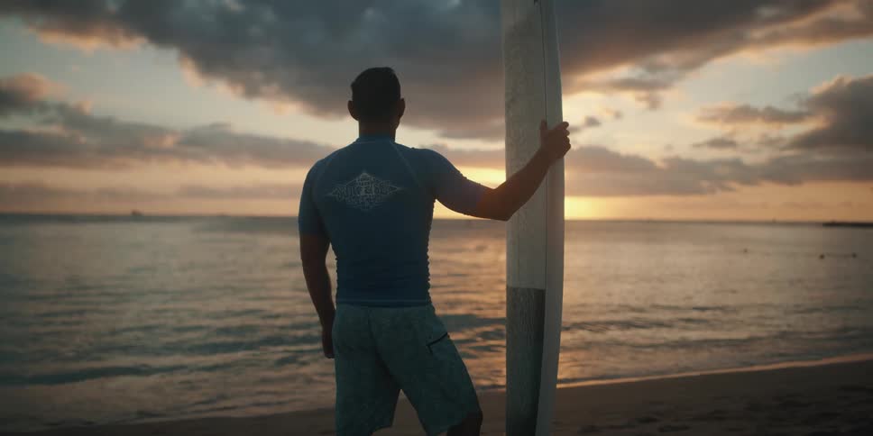 A man standing holding a surfboard looking at the ocean and the sunset
