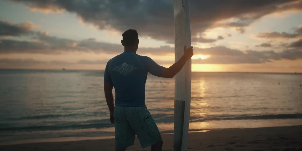 Man holding a surfboard stands on the sand beach and watches sunset