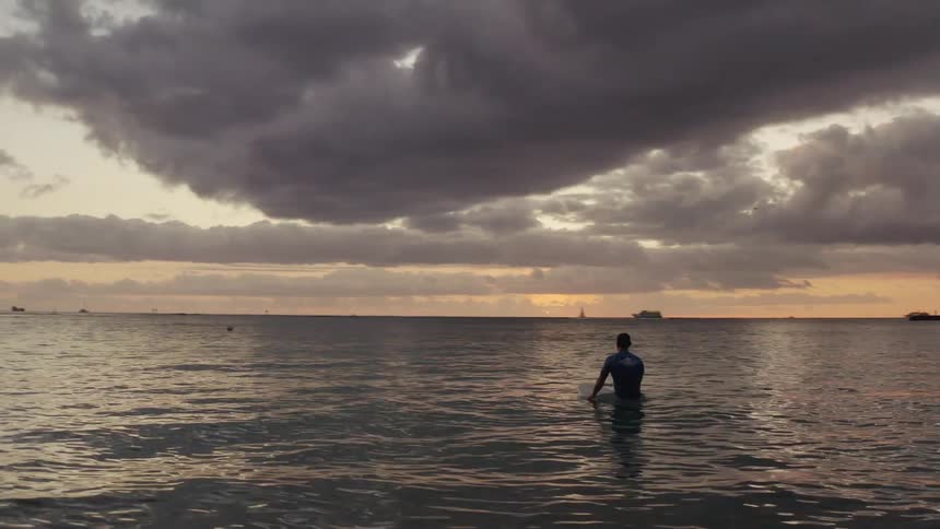Surfer floats on sea over surfboard looking at sunset