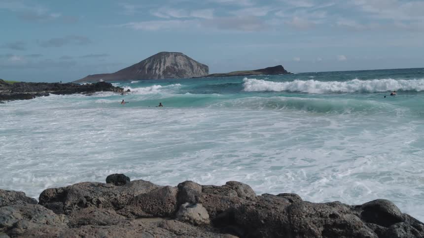 People swim in bright blue ocean with large waves near rocky beach