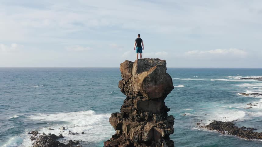 Man stands on top of a rocky cliff and looks out at the ocean