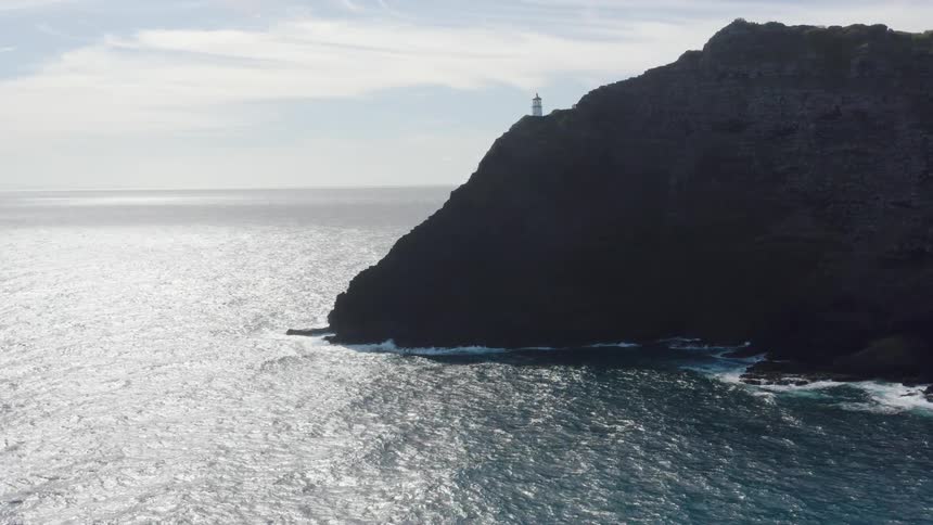 A lighthouse stands on a ridge overlooking the ocean as the sunlight reflects on the surface of the water