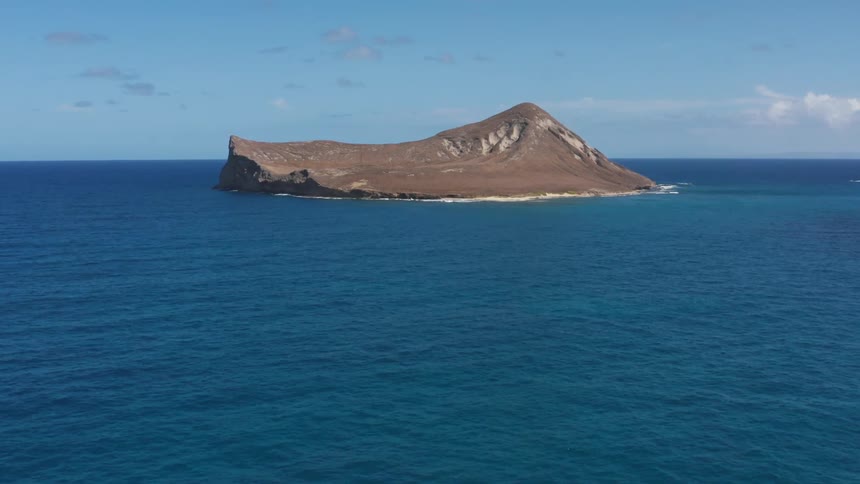 Stony island rices out of azure blue sea under a lightly clouded sky