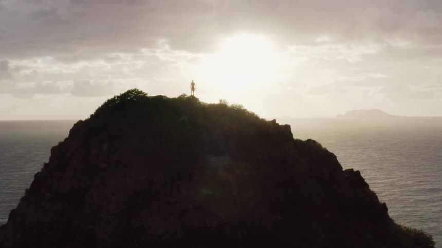 The silhouette of a man standing tall on top of a rocky hill overlooking the ocean