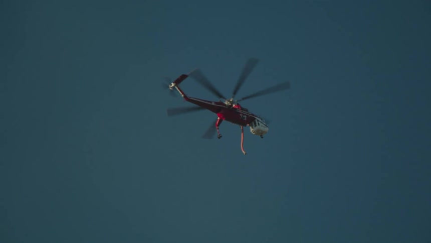 Helicopter flies across clear blue sky trailing a hose