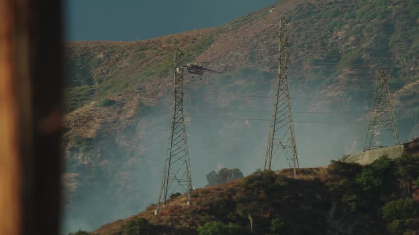 A firefighting helicopter flies over hills as smoke rises from the brush
