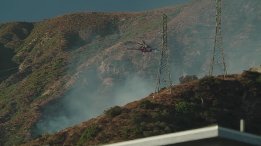 Helicopter flies through smoke rising from mountain near building