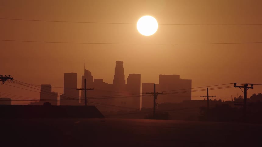 High sun turns sky orange over city downtown as car drives over hill