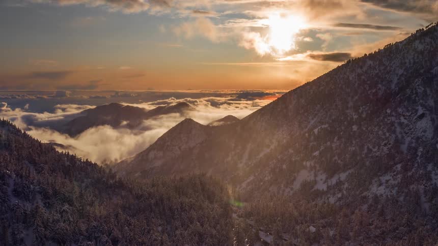 Fog rolls across snow and conifer forest covered mountains under an orange tinted sky