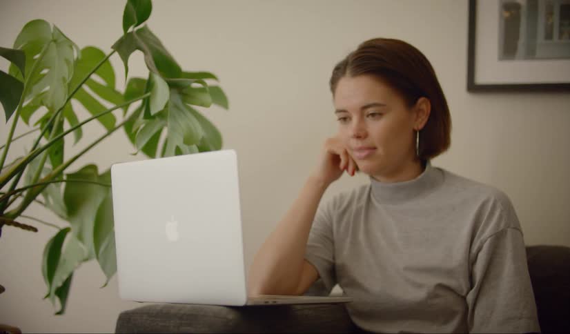 A woman participates in a video conference meeting from home sitting on the couch with her laptop on the arm of the sofa
