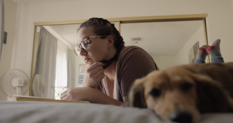 A woman lies on her bed leaning on her arms with her feet up and tapping reading a book with her dog beside her