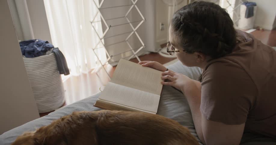 A woman lying on her stomach on her bed reading a book with her small dog sleeping beside her