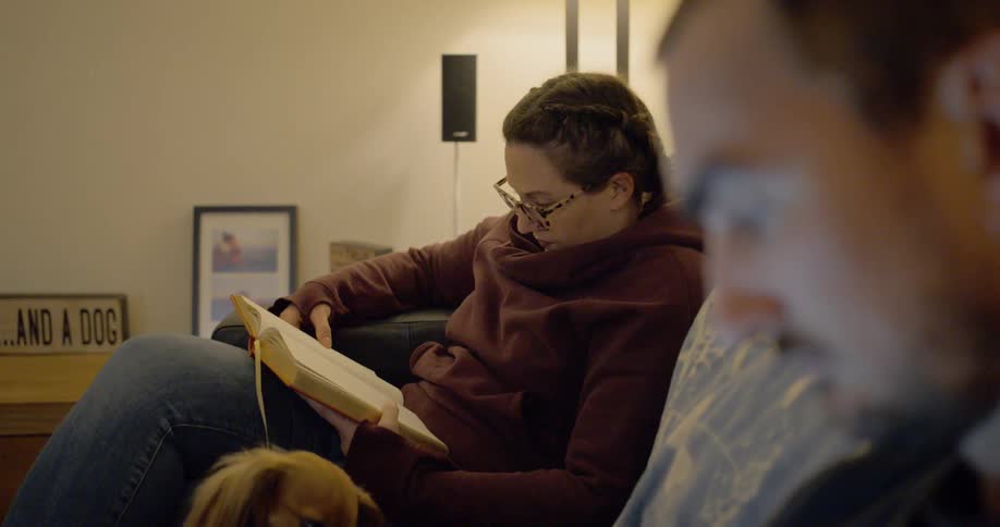 A woman sits on the couch reading with a dog and man