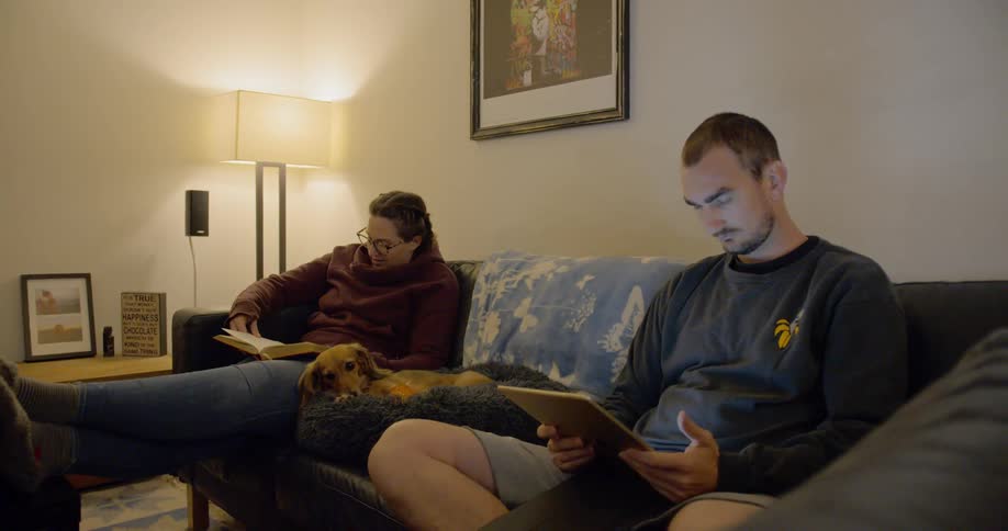 A man and woman sit and read on a couch with a dog between them