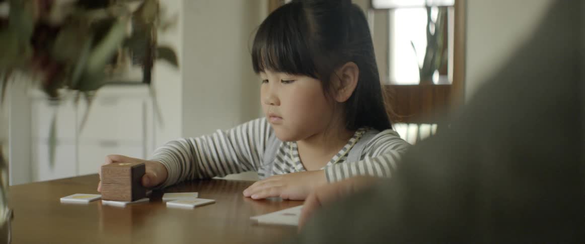 A young girl sits at the dining room table concentrating on a learning game while being instructed