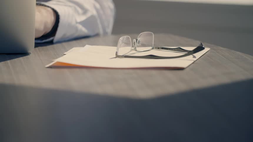 Glasses sit on a folder on a table next to a doctor that is typing on a laptop