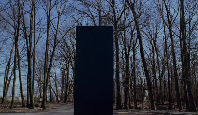 A tall black box sits in the parking lot in front of a walking trail