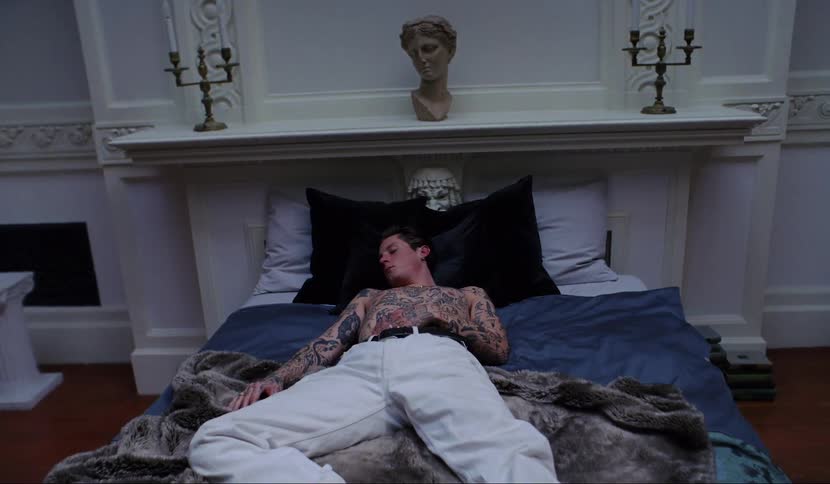 A shirtless tattooed man sleeps on a bed in an ornate bedroom