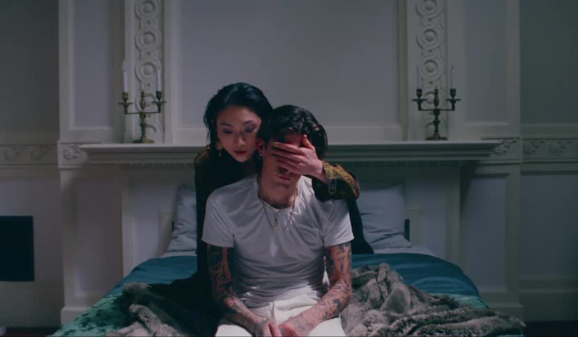 A woman puts her hands over a mans eyes while sitting on a bed