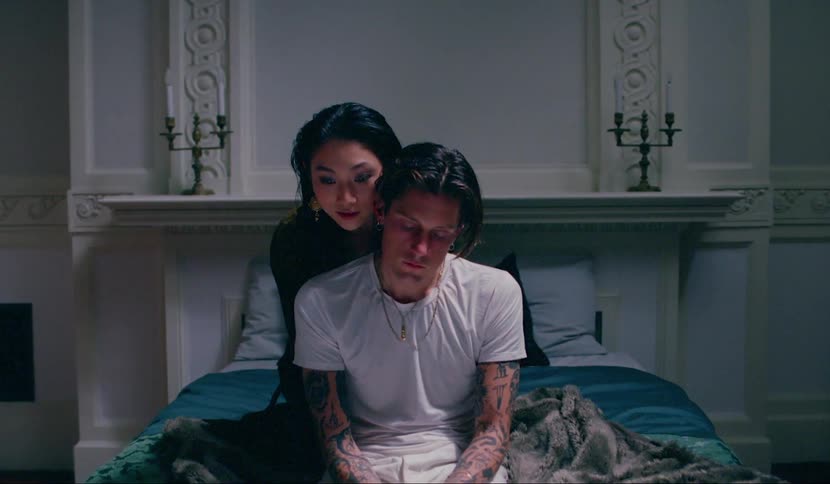 A man sitting on a bed while a woman kneels behind him whispering in his ear