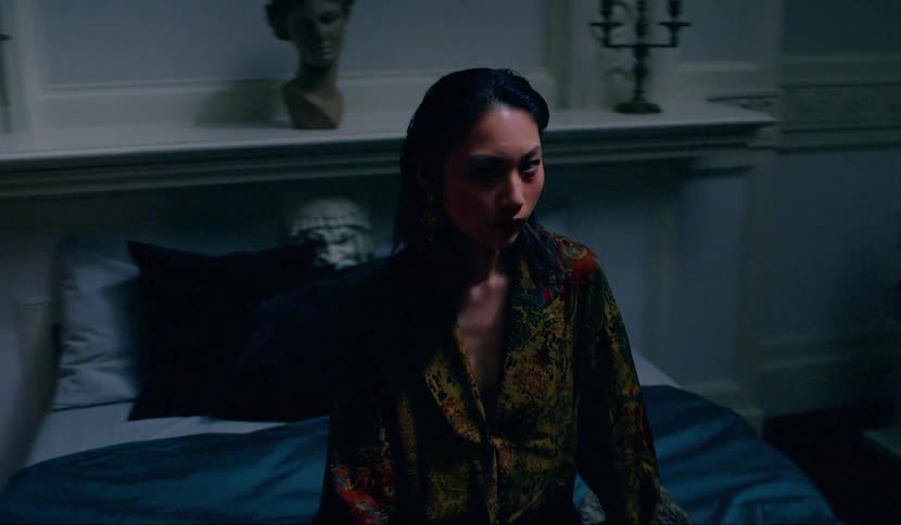A woman sits on a bed in an ornate bedroom tracking someone with her eyes