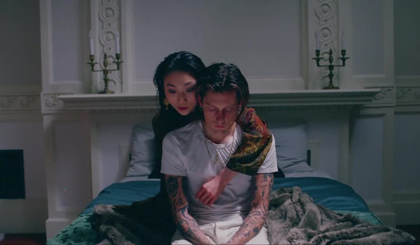 A woman puts her arm around a man from behind while both are on a bed and she whispers in his ear