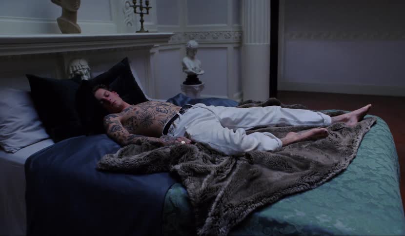 A tattoo covered shirtless man sleeps on a bed in a luxurious room