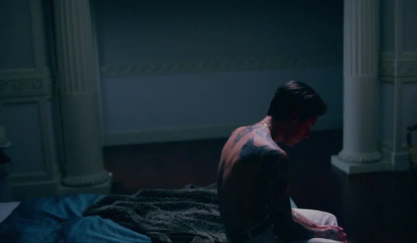 A shirtless man with tattoos sits on the end of a bed looking down at his hands