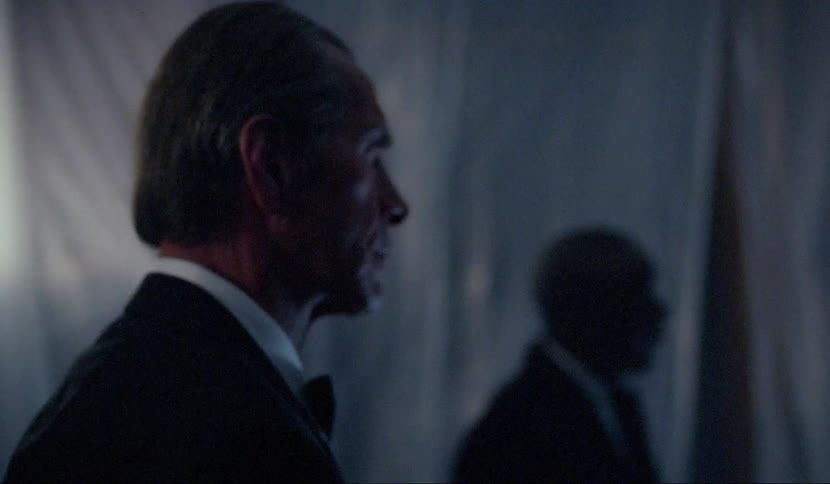 Men stand still in a shadowy room while one other man moves