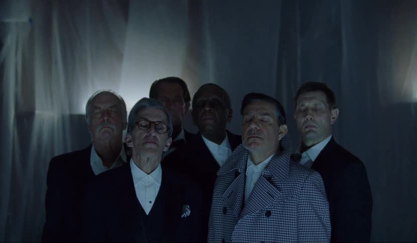 A group of six men stand together with their eyes closed