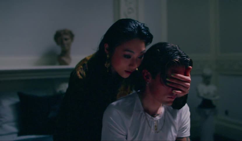 A man sits on a bed as a woman sits behind him and covers his eyes