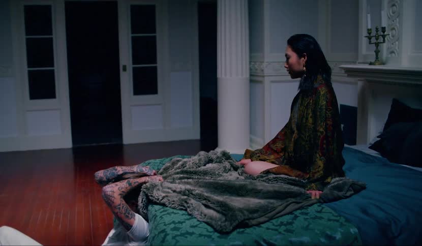 A woman sits on a bed while a man sits on the floor holding the bedspread