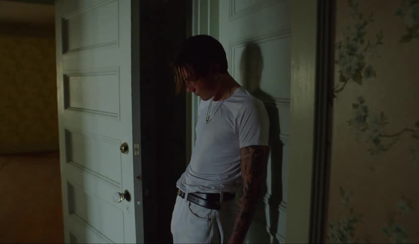 A young white man leans against a wooden door inside a house