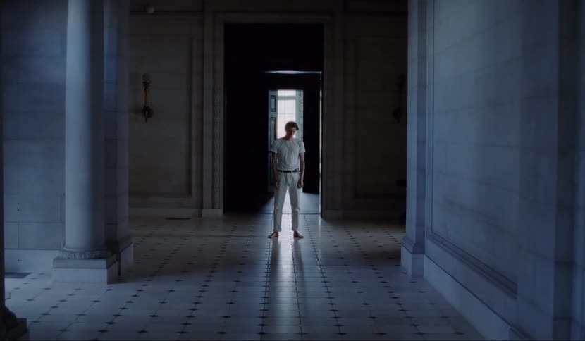 A man wearing a white shirt and pants stands still at the end of a long hallway