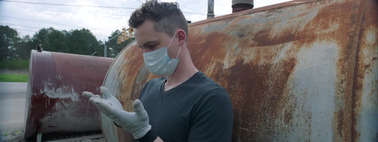 Man sits on ground outdoors near rusted tank examining medical gloves