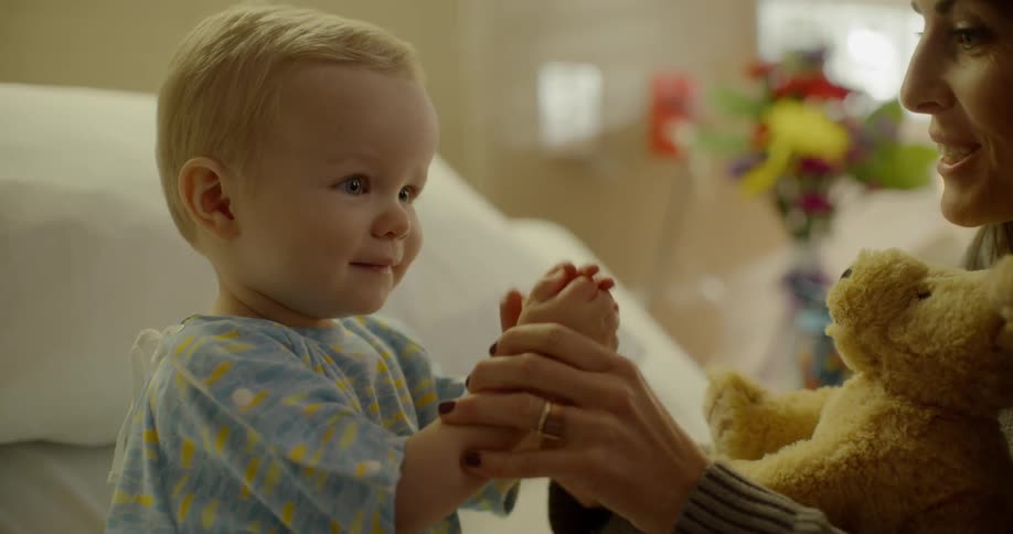A woman with a teddy bear playing with a baby boy in a hospital bed
