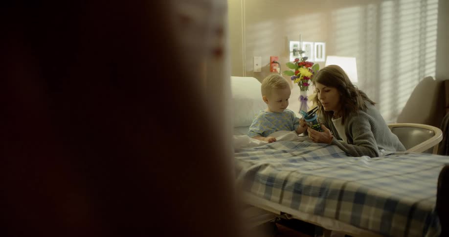 A woman playing with a boy in a hospital bed as a man walks by