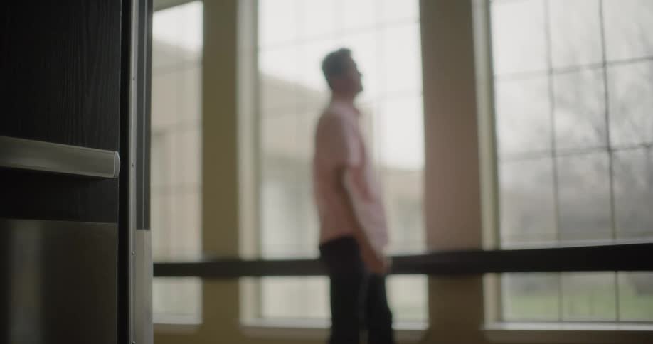 A man looks out a window with his head bowed and walks away