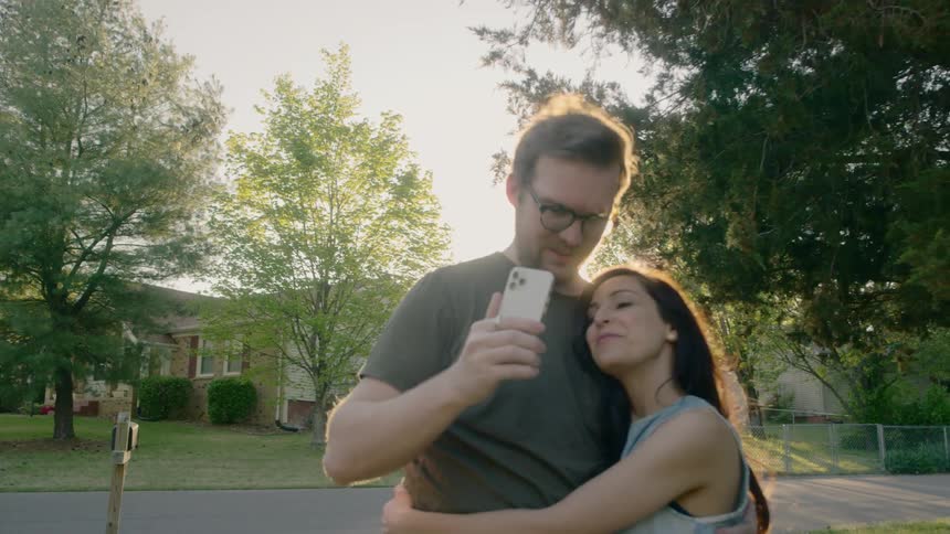 A man and woman filming themselves with a phone while hugging and smiling outdoors